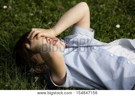 Little boy covering his eyes with his hands. Boy lying on the lawn and he is sad. The youngster screens the eyes from the sun. Caucasian baby in vogue shirt and white shirt. The boy thinks.