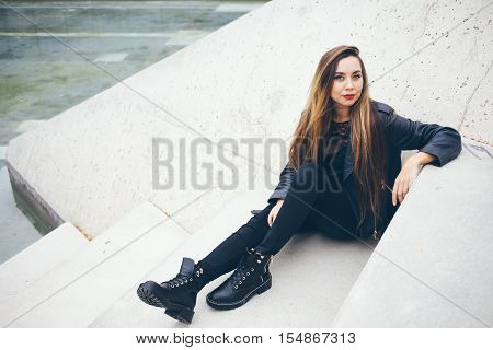 Beautuful young teenage girl is sitting on the concrete steps. Student in alternative style clothes - black leather jacket and big boots. Copy-space area for your advertisement text or design