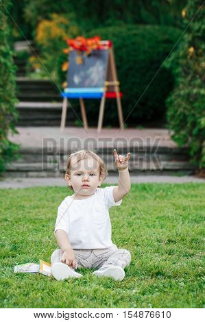 Portrait of funny white Caucasian toddler child kid boy sitting on grass ground outside in summer autumn park by drawing easel with books showing rock metal salute sign back to school