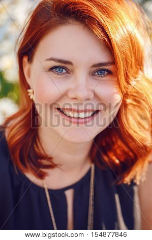 Closeup portrait of smiling middle aged white caucasian woman with waved curly red hair in black dress looking in camera outside in park garden beauty fashion lifestyle concept