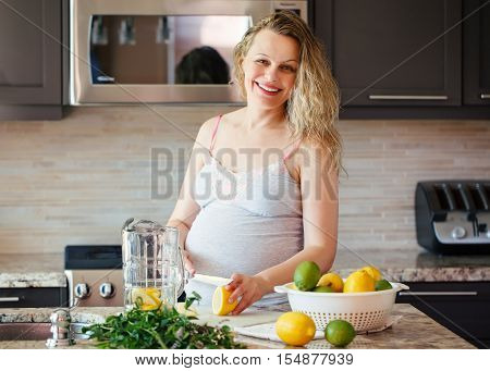 Portrait of smiling white Caucasian blonde pregnant woman cutting citrus lime lemon making juice standing in kitchen at home looking in camera healthy lifestyle concept
