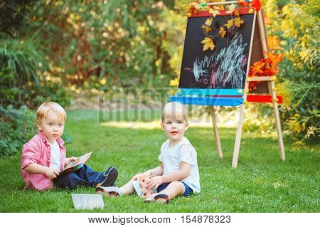 Group of two white Caucasian toddlers children kids, boy and girl sitting in grass outside by drawing easel with books reading studying learning, back to school concept, summer autumn