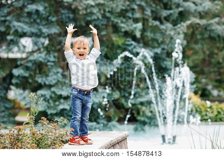 Portrait of cute adorable funny Caucasian little boy toddler in white shirt and blue jeans playing laughing smiling having fun by fountain in summer outside