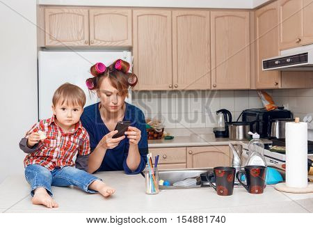 Busy white Caucasian young woman mother housewife with hair-curlers in her hair taking pictures of things objects utensils in her kitchen her kid child son boy sitting beside her eating a cookie smiling and playing crazy busy life