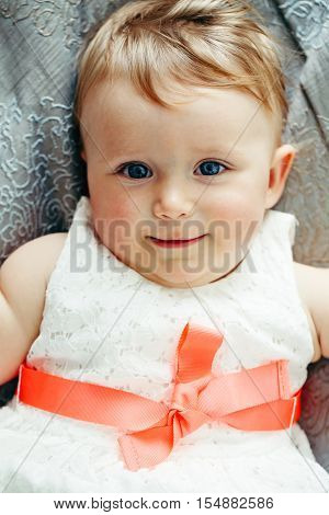 Portrait of cute adorable Caucasian smiling laughing baby girl with blue eyes in white dress with red bow lying on bed looking in camera shot from above overhead top view