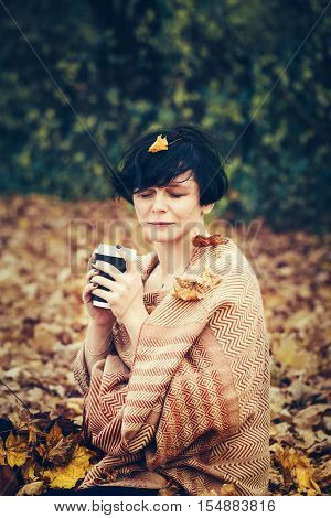 Portrait of middle aged caucasian brunette woman with leaves in her short bob hair yellow orange woolen scarf on her body sitting on groundoutside in park drinking coffee with her eyes closed cozy autumn fall mood feeling concept