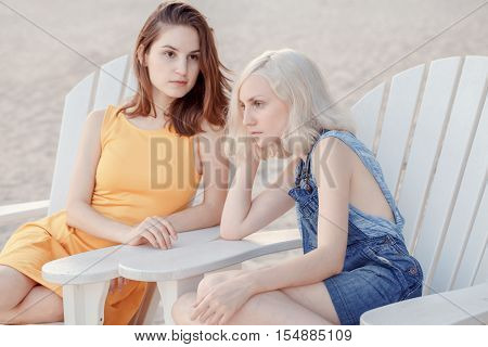 Portrait of two pensive sad white Caucasian unformal young girls hipster students friends outside on beach summer day sitting on chairs silent best friends forever selective focus