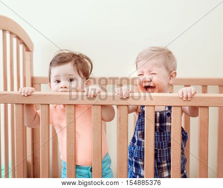 Portrait of two cute adorable funny babies siblings friends of nine months standing in bed crib chewing eating sucking wooden sides crying looking in camera away lifestyle everyday sweet candid moment