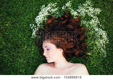 Closeup portrait of young beautiful girl woman with red brown hair lying on grass with white small flowers around her head. View from above top overhead. Concept of spring summer youth happiness