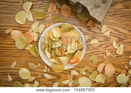 Closeup view from above of colorful veggie potato tomato spinach chips on a white plate and brown paper bag with greesy spots stains on a wooden table