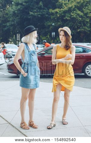 Portrait of two white Caucasian unformal young girls hipster students teenagers friends in dresses hats outside standing talking in urban city street crowded with cars