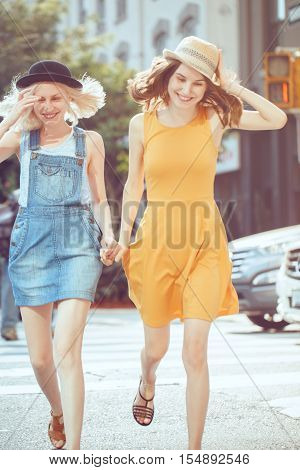 Portrait of two white Caucasian unformal young girls hipster students teenagers friends in dresses hats outside running crossing urban city street crowded with cars best friends forever