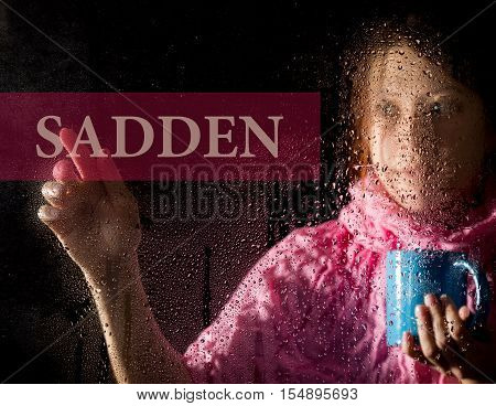 sadden written on virtual screen. young woman melancholy and sad at the window in the rain, she holding a cup of hot coffee or tea.