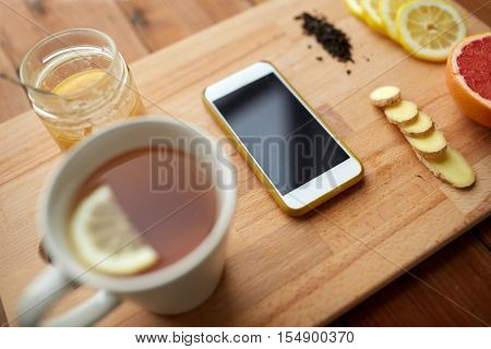 health, traditional medicine, folk remedy and ethnoscience concept - smartphone with cup of ginger tea, honey and citrus on wooden board