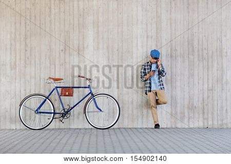 people, style, technology, leisure and lifestyle - young hipster man in earphones with smartphone and fixed gear bike listening to music at city street wall