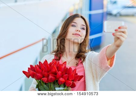 Young Blond Woman With A Bouquet Of Red Tulips Is Taking Selfie On Her Phone.