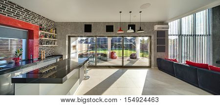 Interior in a modern style with a kitchen zone with a bar, concrete and brick walls, large windows and a door. On the right there is dark sofa with red pillows. Outside there is a terrace and a lawn.