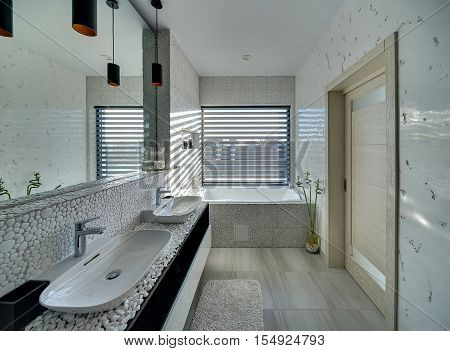 Light bathroom in a modern style decorated with the pebble. There is a bath, two sinks with faucets, big mirror, plant in the pot, window with blinds and a door. On the floor there is a bath carpet.