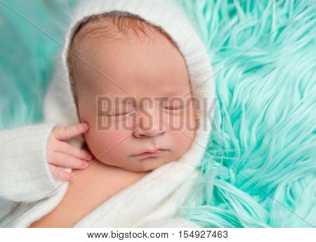 lovely dreamy newborn boy in white romper with hat on fluffy turquoise blanket, close up