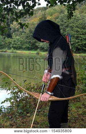 Medieval archer with black hood with the curve span before a lake and looks down