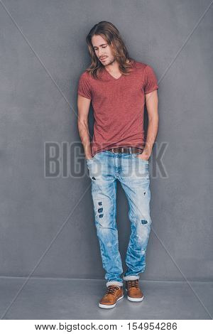 Casually handsome. Full length of handsome young man in casual wear holding hands in pockets and looking thoughtful while standing against grey background