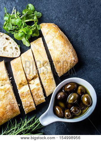 Bread ciabatta, arugula, olives, rosemary on stone slate black background. Top view