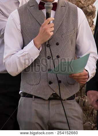 A Happy Groom In A Gray Fashion Vest With A Red Butterfly Holds A Microphone And A Piece Of Paper Wi