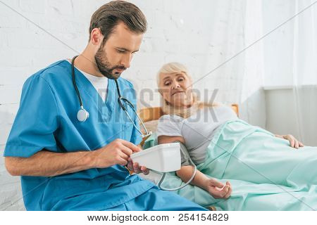 Young Male Nurse With Stethoscope Holding Blood Pressure Monitor While Measuring Blood Pressure To S