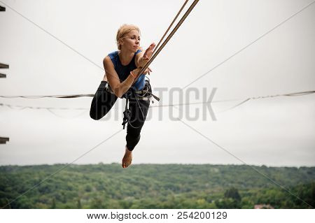 Beautiful Blonde Woman Balancing On A Slackline Against The Background Of Grey Sky And Trees