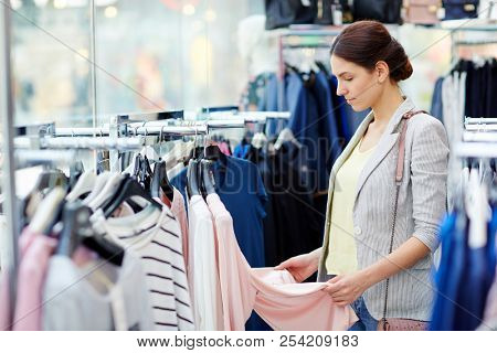 Side view of attractive young woman standing near rail and looking at nice apparel while sending time in clothes shop