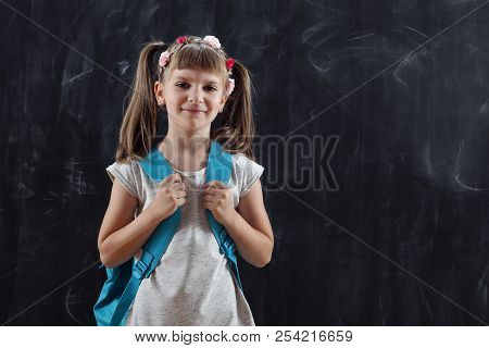 Schoolgirl Wearing Backpack, Standing In Front Of A Blackboard On Her First Day Of School