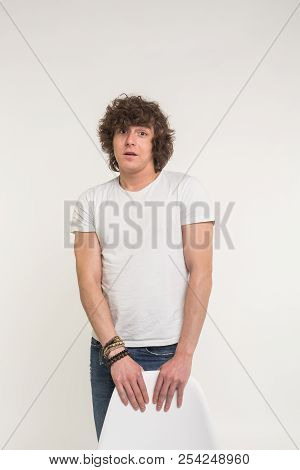 Handsome Young Man With Curly Hair Standing In White T-shirt Behind A Chair On White Background