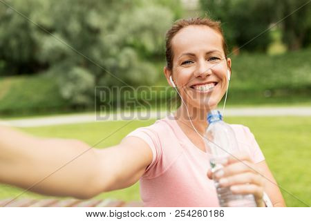 fitness, sport and healthy lifestyle concept - happy woman in earphones and water taking selfie in park