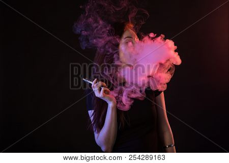 Young Woman In Neon Light And Smoke Of E-cigarettes Or Vape On Black Background