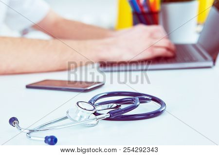 Stethoscope With Smartphone And Laptop On The Desk. Male Doctor Working In Hospital Writing A Prescr