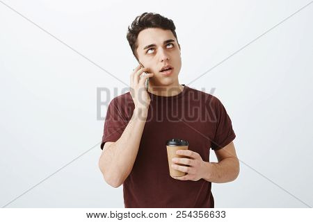 Portrait Of Annoyed Pissed European Guy In Red T-shirt, Holding Cup Of Coffee, Talking On Smartphone