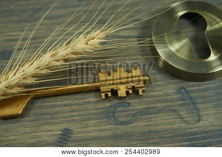Conceptual Close-up Of A Vintage Golden Key And Ear Of Wheat On An Old Wooden Table As Symbol For Ac