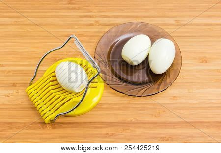 Manual Egg Slicer With Peeled Egg In It And Two Boiled Peeled Eggs On A Dark Glass Saucer On A Bambo