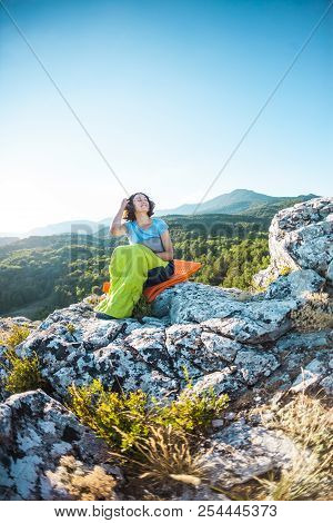 A Woman Is Sitting In A Sleeping Bag At Sunset. Camping On Top Of A Mountain. A Smiling Girl Rests A