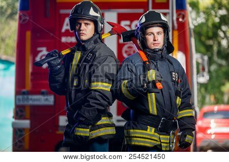 Photo of two firemen with axes in hands near fire engine