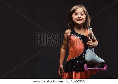Joyful Little Girl Dressed In Skater Dress Holds Ice Skates. Isolated On A Dark Textured Background.