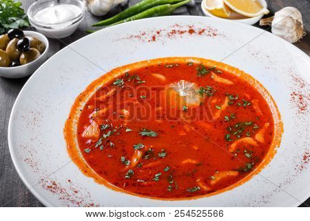 Soup With Meat, Olives, Herbs And Lemon In Plate, Served With Sour Cream, Peppers On Dark Wooden Bac