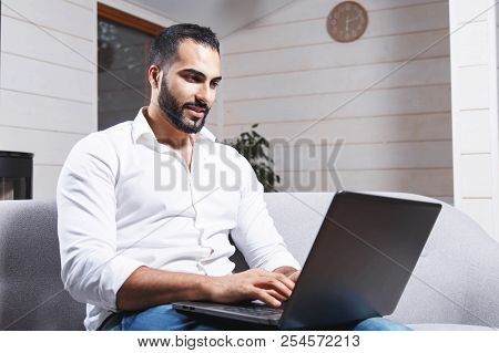 Handsome Young Enterpreneur, Bearded Man Wears White Shirt Sitting On The Sofa And Working With Lapt