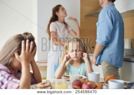 Two upset little girls sitting by served table and feeling anxious about their parents argument on background