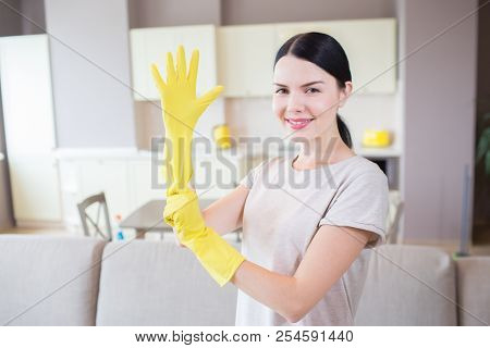Attractive Woman Looks On Camera. She Stands In Studio Apartment And Puts On Yellow Glove On Right H