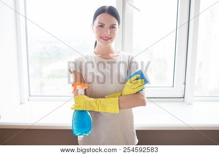 Happy And Positive Housewife Stands In Front Of Window And Poses. She Looks On Camera. Girl Keeps He