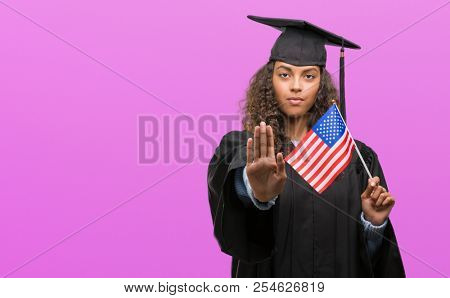Young hispanic woman wearing graduation uniform holding flag of United States with open hand doing stop sign with serious and confident expression, defense gesture