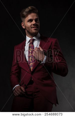 portrait of smiling man arranging his grena suit collar looking to side while sitting on grey wallpaper background
