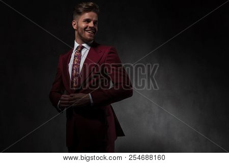 portrait of joyful young man unbuttoning his red suit and looking to side while standing on grey wallpaper background