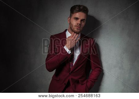 portrait of sensual elegant man in grena suit touching his neck while standing on grey wallpaper background with a hand in pocket